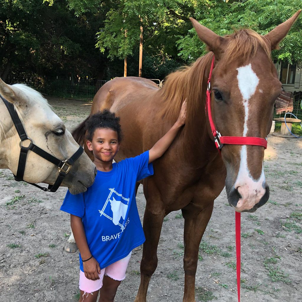 Young girl stands between two horses with her arm reaching up to pet one of the horses. Image from Bravehearts Equine Therapy.